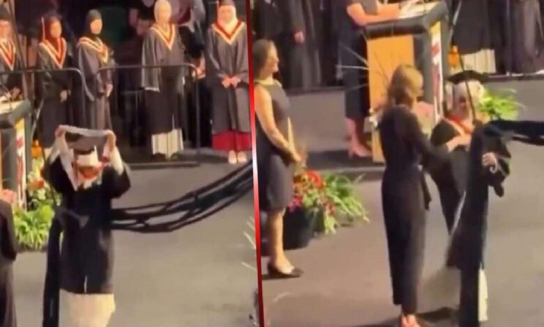 Graduate in cap and gown raises the hood on stage during a graduation ceremony, with other graduates and a speaker at a podium in the background.