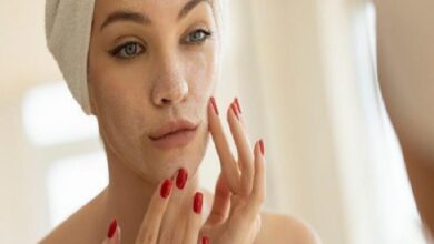 Woman with a towel on her head applies skincare to her face in front of a mirror, checking her skin.