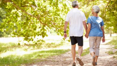 Older couple walks hand in hand along a sunlit park path lined with trees