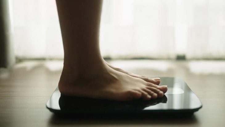 Bare feet standing on a glass-top bathroom scale on a wooden floor in a bright room
